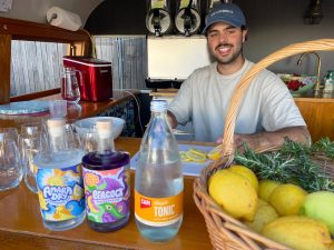 Amara Dry and Peacock Gin with their vibrant labels stand next to a bottle of Capi Tonic and a basket of Lemons. A man with a moustache and a cap on is in the background